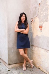 Kristin standing in front of an outdoor wall.  Arms folded, Ankles crossed.