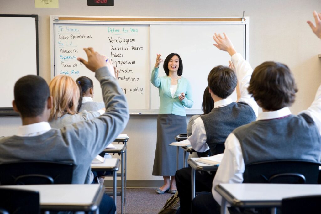 teacher in front of a classroom of high school students raising hands