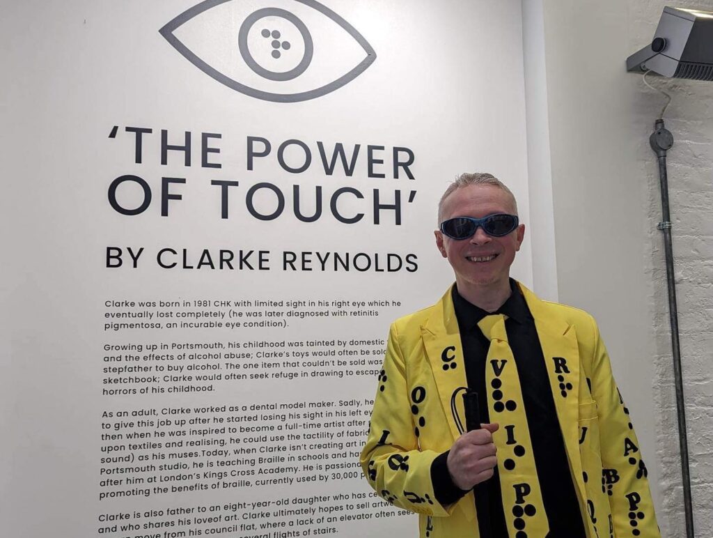 Clarke Reynolds smiling wearing his signature yellow and black braille suit standing in front of his art exhibit titled"The Power of Touch"
