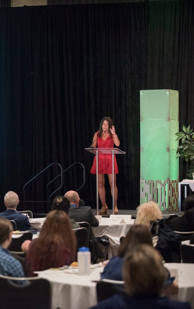 Kristin Smedley delivering a keynote from the podium to a full conference ballroom