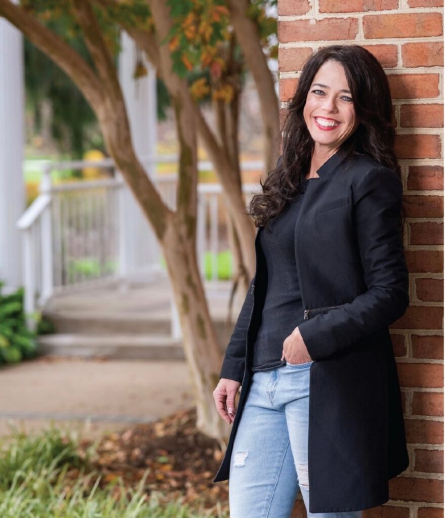 Kristin Smedley smiling, leaning against a brick wall in a black jacket and jeans