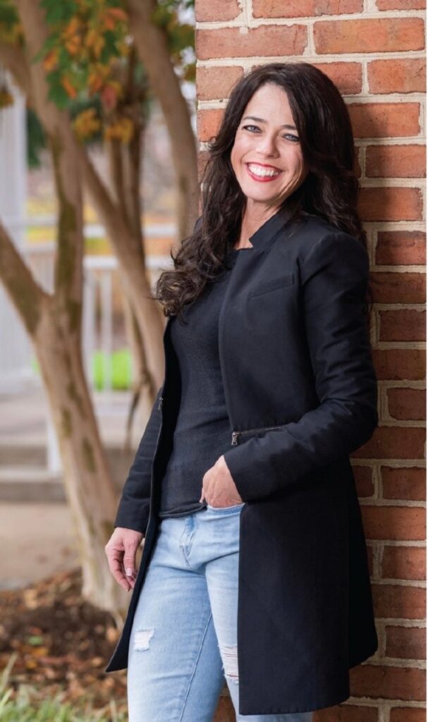Kristin Smedley smiling, leaning against a brick wall in a black jacket and jeans